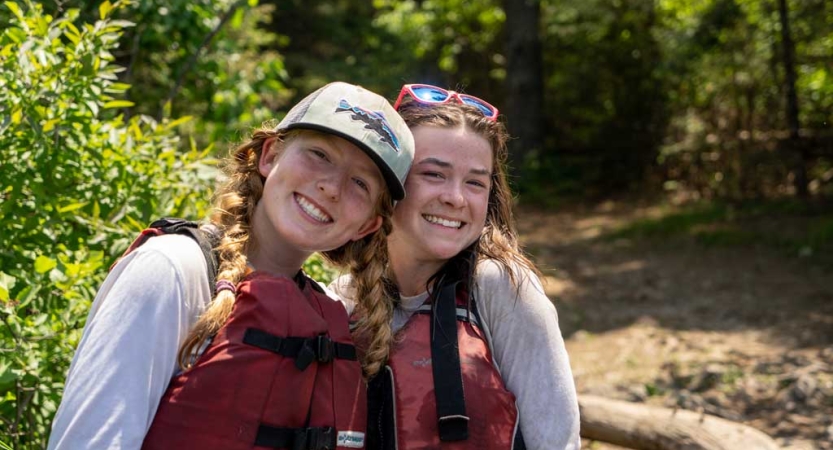 Two young people wearing life jackets stand in a wooded area and smile for the photo.